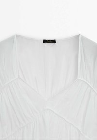 Blouse blanche avec un décolleté en V, texture froncée et corsage ajusté. Présente une petite étiquette noire au cou avec le nom de la marque.