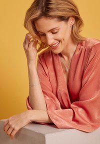 Korallfärgad satinblus med vida ärmar, silversmycken inklusive ett delikat armband och halsband, mot en gul bakgrund.