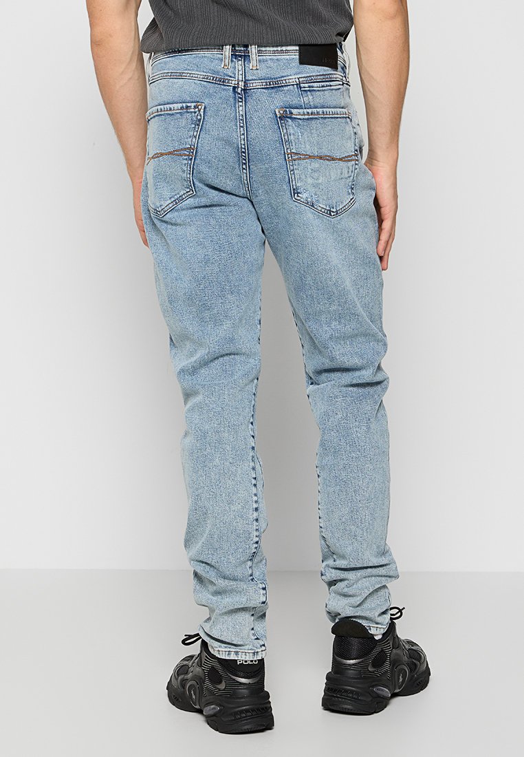 Mann in hellblauen Jeans und schwarzen Sneakers, stehend mit entspannten Händen an den Seiten, vor einem einfarbigen hellgrauen Hintergrund.