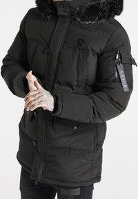 Chaqueta acolchada negra con capucha de peluche sintético, que cuenta con múltiples bolsillos, detalle de cremallera en la manga y parche con logo en el pecho. Textura acolchada y ajuste ceñido.