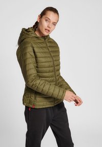 Olivgrüne gefütterte Jacke mit Kapuze, Frontreißverschluss und seitlichen Taschen; mit horizontaler Steppung und glatter Textur. Kombiniert mit schwarzen Hosen.