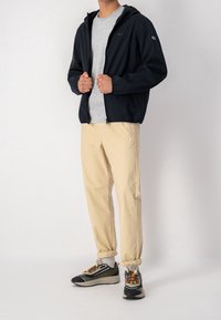 Marinblå zip-up jacka med huva, grå t-shirt, ljusbeiga byxor och svarta sneakers med färgglada detaljer och texturerade sålar.