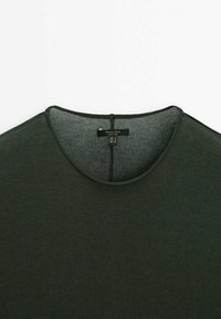 Mørkegrøn crew neck sweater med synlig indvendig søm og et tøjmærke ved halsåbningen, lagt fladt på en hvid baggrund.