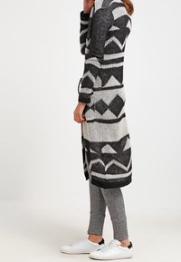 Žena v dlouhém kardiganu s geometrickým vzorem v černé a bílé barvě, šedých legínách a bílých teniskách stojící bokem.