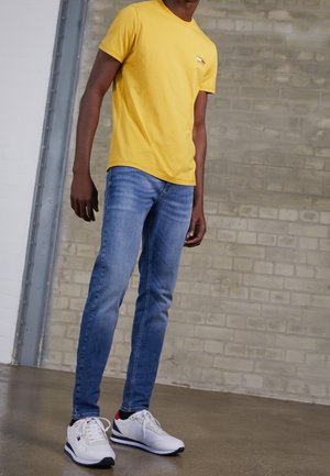 Person wearing a yellow t-shirt, blue slim-fit jeans, and white sneakers standing indoors against a gray brick wall background.