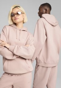 Lysrosa hoodie og sæt af sweatpants i blødt stof, der har hætte, lommer og broderet logo på brystet og benet. Modellen poser med solbriller.