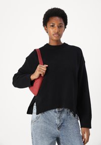 Črna pletena jopica z rebrastimi manšeti in širokim ovratnikom, v kombinaciji s svetlo modrimi hlačami z visokim pasom in majhno rdečo usnjeno torbico čez ramo.