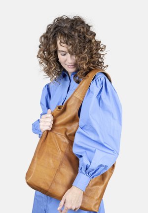 Brun læder skuldertaske med en blød tekstur, der har en enkelt skulderrem og et minimalistisk design. Den blå bluse båret af modellen tilføjer kontrast.