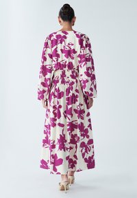 Robe longue crème avec imprimé floral violet, manches larges et taille élastique au dos. Tissu lisse et texturé avec une silhouette fluide.
