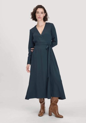 Teal wikkeljurk met lange mouwen, een diepe V-hals en een gestrikt taille. Gepaard met kniehoge, bruine leren haklaarzen. Gladde textuur.