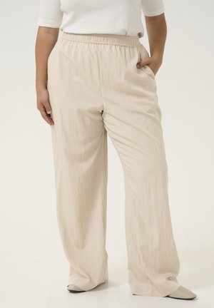 Personne portant un pantalon beige ample avec une ceinture élastique et des chaussures blanches, debout avec une main dans la poche sur un fond uni.
