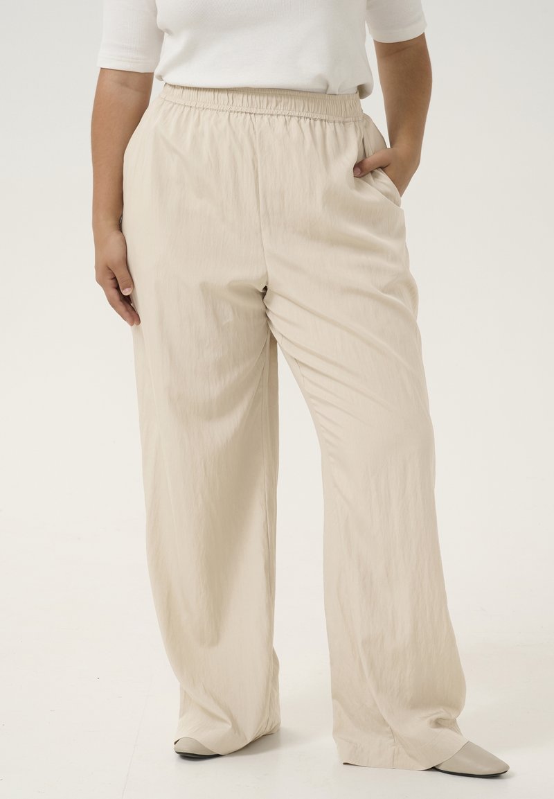 Personne portant un pantalon beige ample avec une ceinture élastique et des chaussures blanches, debout avec une main dans la poche sur un fond uni.