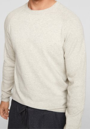 Mann trägt cremefarbenen, langärmeligen, strukturierten Pullover und dunkle Hose mit Kordelzug vor einfachem Hintergrund, Oberkörper sichtbar.