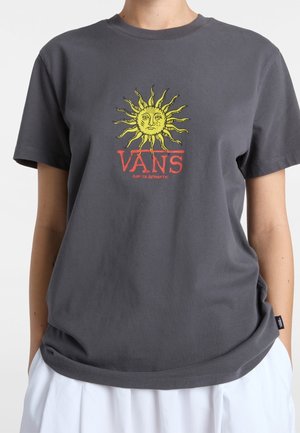 Osoba nosząca ciemnoszarą koszulkę Vans z żółtą grafiką słońca i czerwonym logo Vans, w połączeniu z białymi spodniami, ręce w kieszeniach.