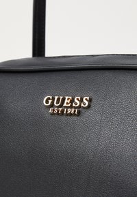 Bolso de mano de imitación cuero negro con una textura suave. Presenta un detalle del logo dorado "GUESS EST 1981" en la parte frontal. Correa ajustable negra.