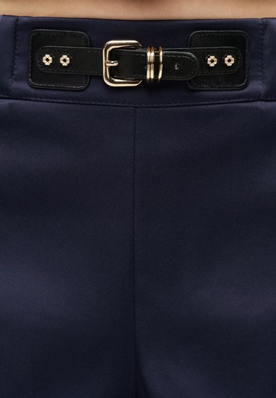 Κοντινό πλάνο από μπλε παντελόνι με μαύρη δερμάτινη ζώνη που διαθέτει χρυσή αγκράφα και δύο διακοσμητικούς χρυσούς δακτυλίους.