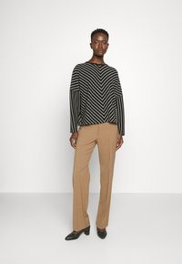 Black and white chevron-patterned sweater with wide sleeves, paired with fitted tan trousers and black heeled shoes. Simple background.