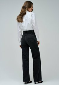 Mujer con cabello largo castaño, vistiendo una camisa blanca de botones, jeans negros de cintura alta y tacones negros, de pie de lado sobre un fondo gris.