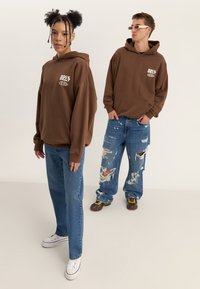 Brun oversized hettegenser med frontlomme, kombinert med blå slitte jeans. Modellen har på seg hvite joggesko, og står på en nøytral bakgrunn.