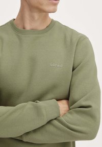 Felpa verde oliva con colletto a girocollo, texture morbida e un sottile logo "blend" dello stesso colore sul petto.