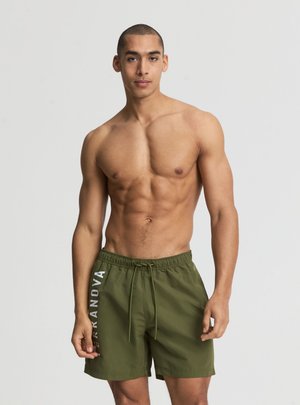 Giovane uomo in forma in piedi, indossa pantaloncini da bagno verde oliva con scritta verticale bianca "TERRANOVA" sulla gamba sinistra, su uno sfondo chiaro uniforme.