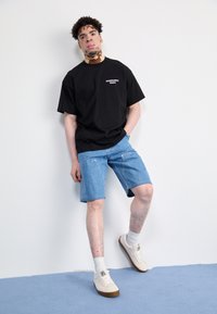 Black t-shirt with white text, light blue denim shorts, white socks, and beige sneakers. Smooth fabric and relaxed fit.