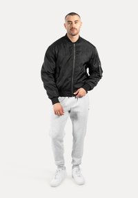 Giubbotto bomber nero con cerniera frontale e polsini a costine, abbinato a pantaloni della tuta grigio chiaro. Il modello è in piedi su uno sfondo neutro.