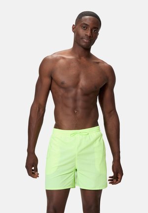 Shirtless man with short hair wearing light green swim shorts against a plain light background, standing and looking at the camera.