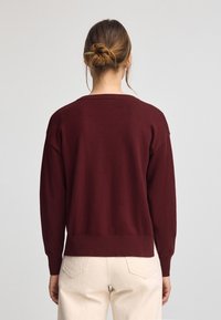 Burgundy stickad tröja med avspänt snitt, sänkta axlar och ribbad nederkant. Bärs över ljusa byxor, vilket visar en mjuk textur.