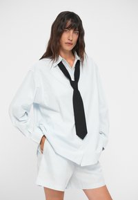 Υπέρμετρα ανοιχτό μπλε ριγέ πουκάμισο με μακριά μανίκια, συνδυασμένο με λευκά σόρτς. Μια μαύρη γραβάτα είναι χαλαρά δεμένη γύρω από τον γιακά.