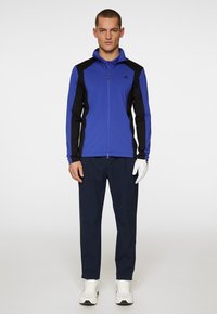 Herrarnas idrottsjacka i blått med svarta detaljer, dragkedja i kragen, lång ärm, kombinerad med marinblå byxor och vita sneakers. Texturerad tyg.