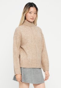 Beige gyapjú keverék pulóver, bordázott garbó nyakkal, lecsúszott vállakkal és szűkített csípőrésszel. Szürke mini szoknyával párosítva.