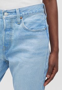 Jean en denim bleu clair porté par une personne à la peau foncée, avec une poche avant, un bouton, une boucle de ceinture et des coutures visibles.
