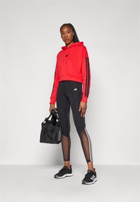 Rode cropped hoodie met zwarte strepen, zwarte leggings met meshpanelen en witte accenten, zwarte sneakers en een zwarte sporttas.