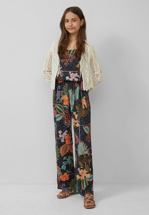 Combinaison noire à motifs floraux avec des couleurs vives et un design à jambes larges, associée à un cardigan en tricot crème. Sandales plates marron visibles.
