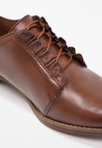 Chaussure en cuir brun avec un bout arrondi, des lacets décoratifs et des coutures sur les côtés. Semelle plate avec une finition légèrement texturée.