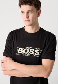 T-shirt en coton noir avec un logo "BOSS" blanc décoloré sur le devant. Manches courtes et col rond. Texture douce.