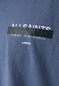 Blå bomulds T-shirt med "ALLSAINTS" i hvid broderi, med et sort bånd, der dækker teksten nedenfor, og "LONDON" trykt i hvid.