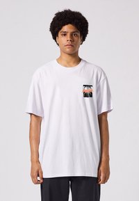 White cotton t-shirt with a relaxed fit, featuring a small black-and-orange graphic on the left chest area and short sleeves.
