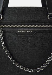 Un sac à main en cuir noir avec une poche zippée et un accent en chaîne argentée. Le nom de la marque "Michael Kors" est affiché en évidence.