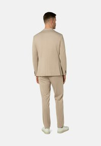 Beige Sakko mit passender Hose, aus glattem Stoff gefertigt. Verfügt über minimale Nähte und einen dezenten Glanz. Weiße Sneaker runden den Look ab.
