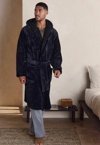 Navy plush bathrobe with a hood, tied at the waist, featuring two front pockets. Model wears light gray striped pants. Textured background.