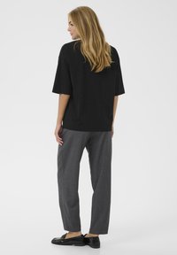 Femeie cu păr lung blond, întoarsă lateral, îmbrăcată cu un top negru lejer cu mâneci scurte, pantaloni gri cu croială dreaptă și mocasini negri, pe fundal alb.