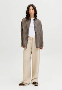 Donna con capelli ricci e scuri, che indossa una camicia a quadri marrone con bottoni, una maglietta bianca corta, pantaloni beige a gamba larga e scarpe marroni scure.