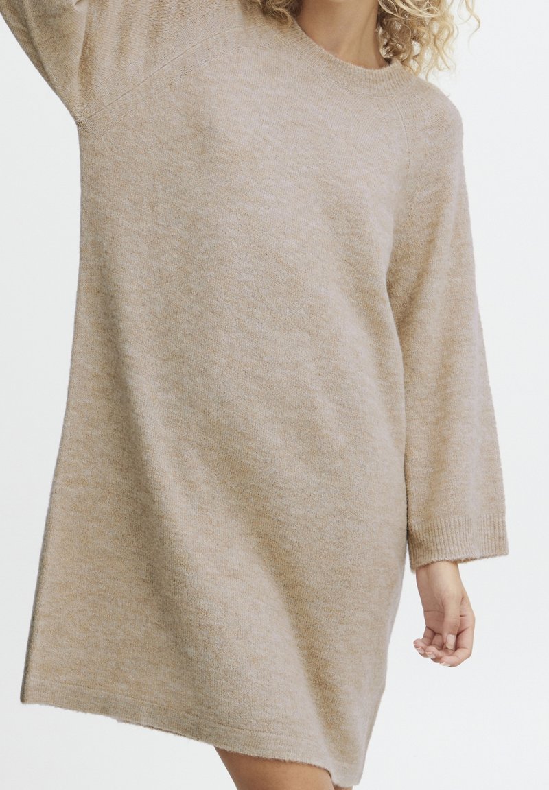 Femme portant une robe pull ample en tricot beige à manches longues, debout devant un fond blanc uni.