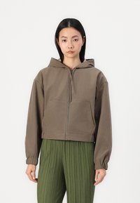 Sudadera marrón de corte corto con cremallera y capucha, que cuenta con dos bolsillos frontales y puños elásticos. Usada sobre pantalones de pliegues verdes, de textura suave.