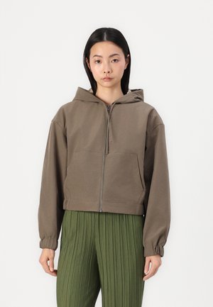 Brun cropped zip-up hettegenser med hette, med to frontlommer og elastiske mansjetter. Båret over grønne plisserte bukser med jevn tekstur.