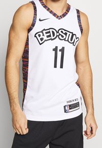 Maillot de basketball blanc sans manches avec "BED-STUY" et le numéro 11 en noir, bordure colorée, et étiquette Brooklyn Nets sur le bas devant.