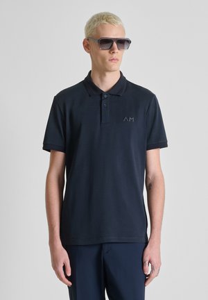 Joven con cabello rubio corto que lleva gafas de sol oscuras, una camiseta polo azul marino con el logo "AM" y pantalones a juego, posando contra un fondo liso.