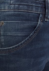 Jeans de denim azul oscuro con costuras visibles, un bolsillo trasero y un acento de botón metálico. La textura muestra un ligero desgaste. Vista en primer plano.